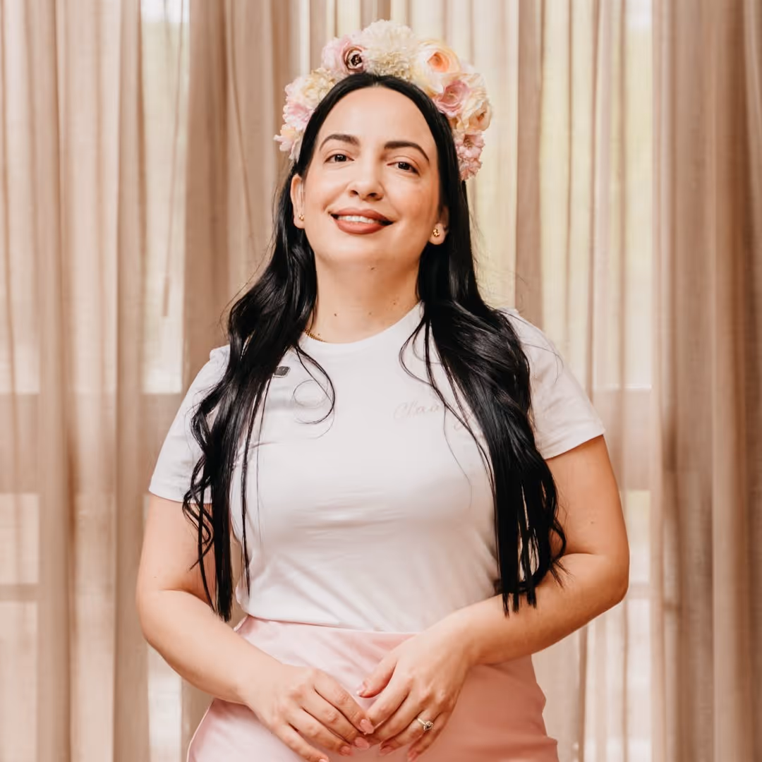 Mujer con cabello largo y oscuro, sonriente, usando una diadema de flores y camiseta blanca frente a cortinas beige.