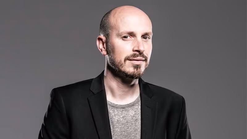 Hombre calvo con barba y chaqueta negra mirando hacia la cámara sobre fondo gris.