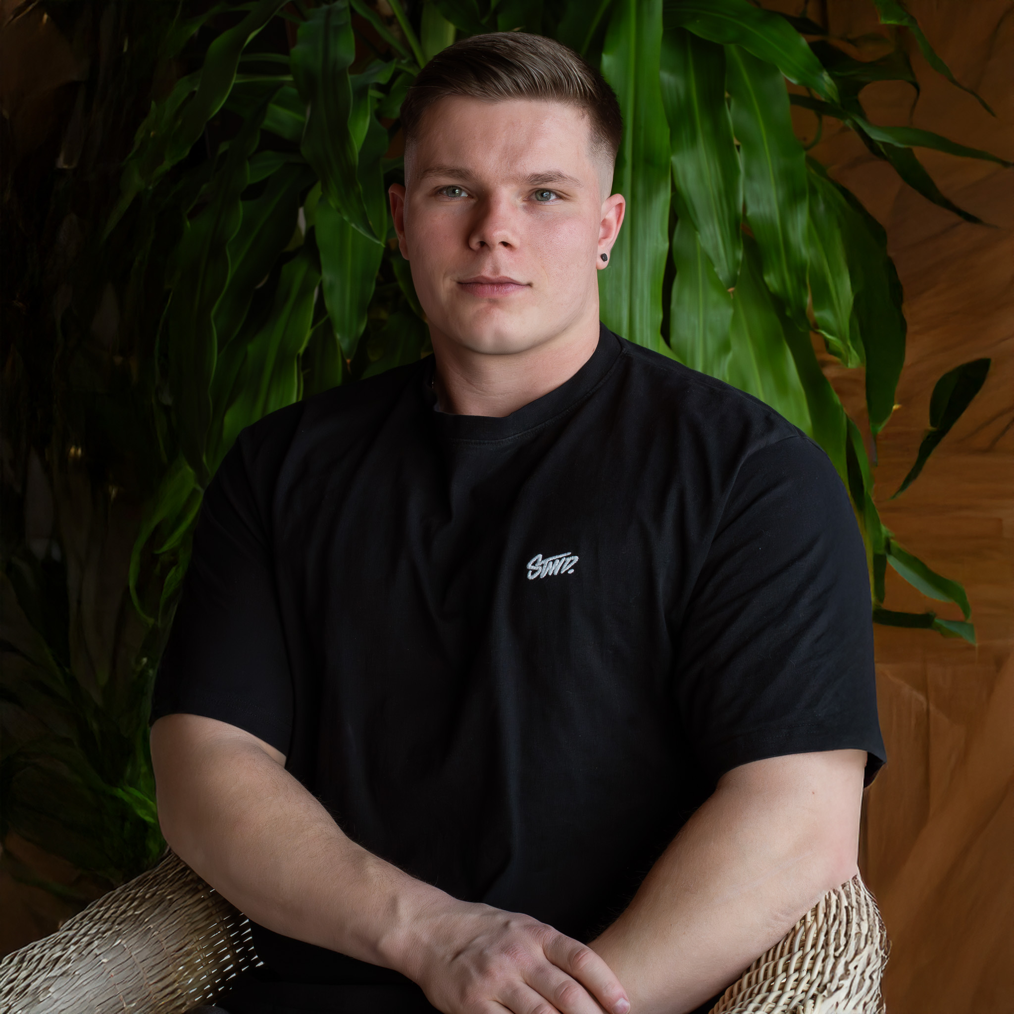 Junger Mann mit kurzem Haar und schwarzem T-Shirt sitzt auf einem Stuhl vor grünen Pflanzen.