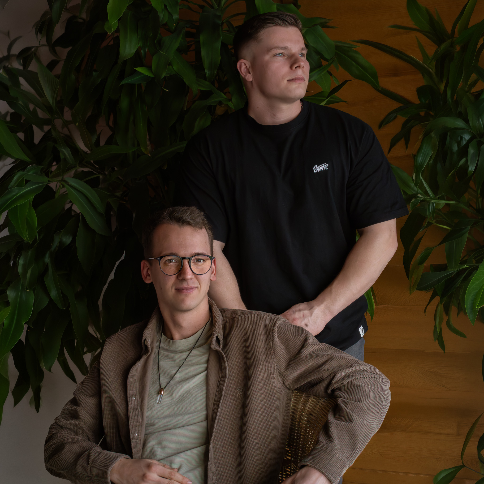 Zwei junge Männer vor einer Holzpaneel-Wand und grünen Pflanzen, einer sitzt mit Brille und brauner Jacke, der andere steht hinter ihm in schwarzem T-Shirt.