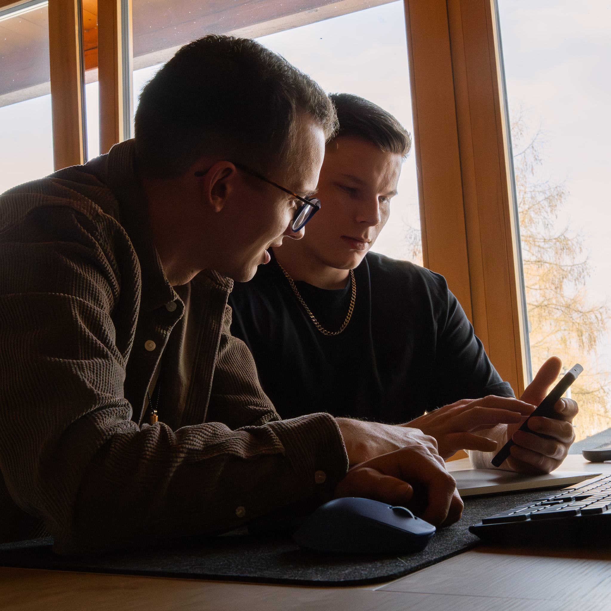 Zwei junge Männer sitzen an einem Schreibtisch vor einem Fenster und schauen auf ein Smartphone.