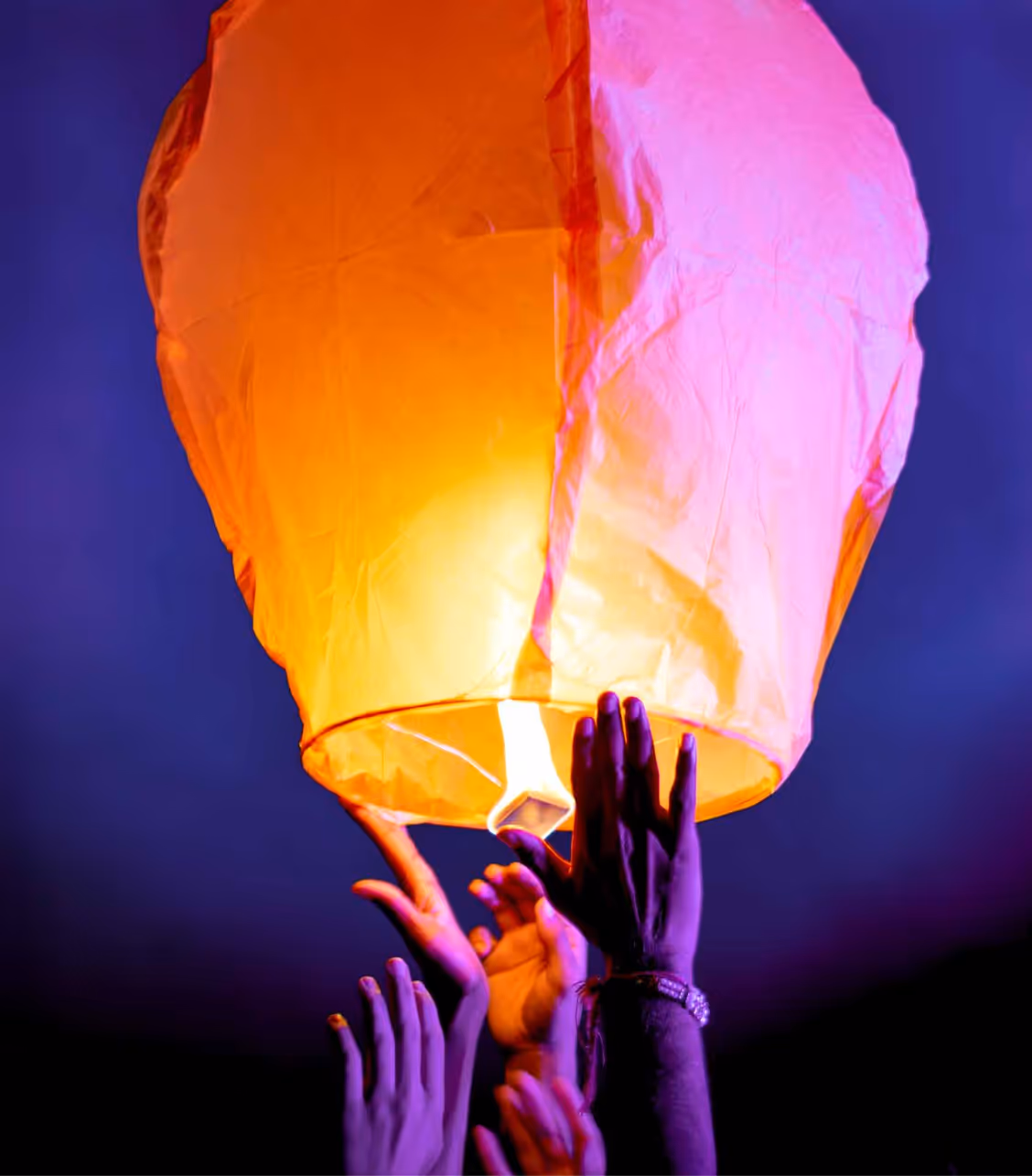 Plusieurs mains relâchant une lanterne céleste orange et rose dans le ciel nocturne.