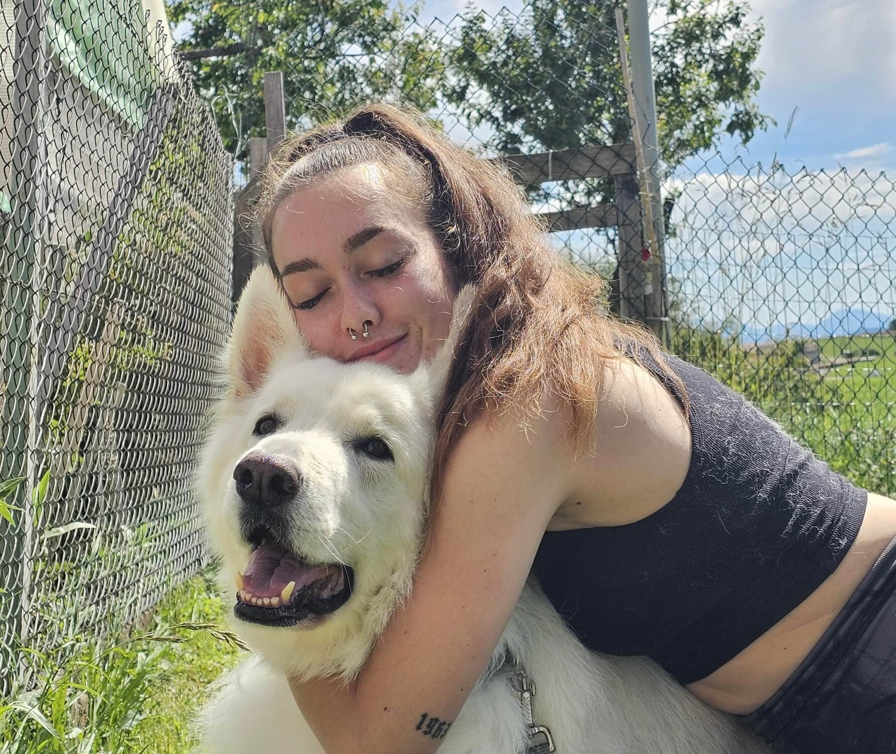 Young woman with long hair hugging a happy white fluffy dog outdoors by a chain-link fence.