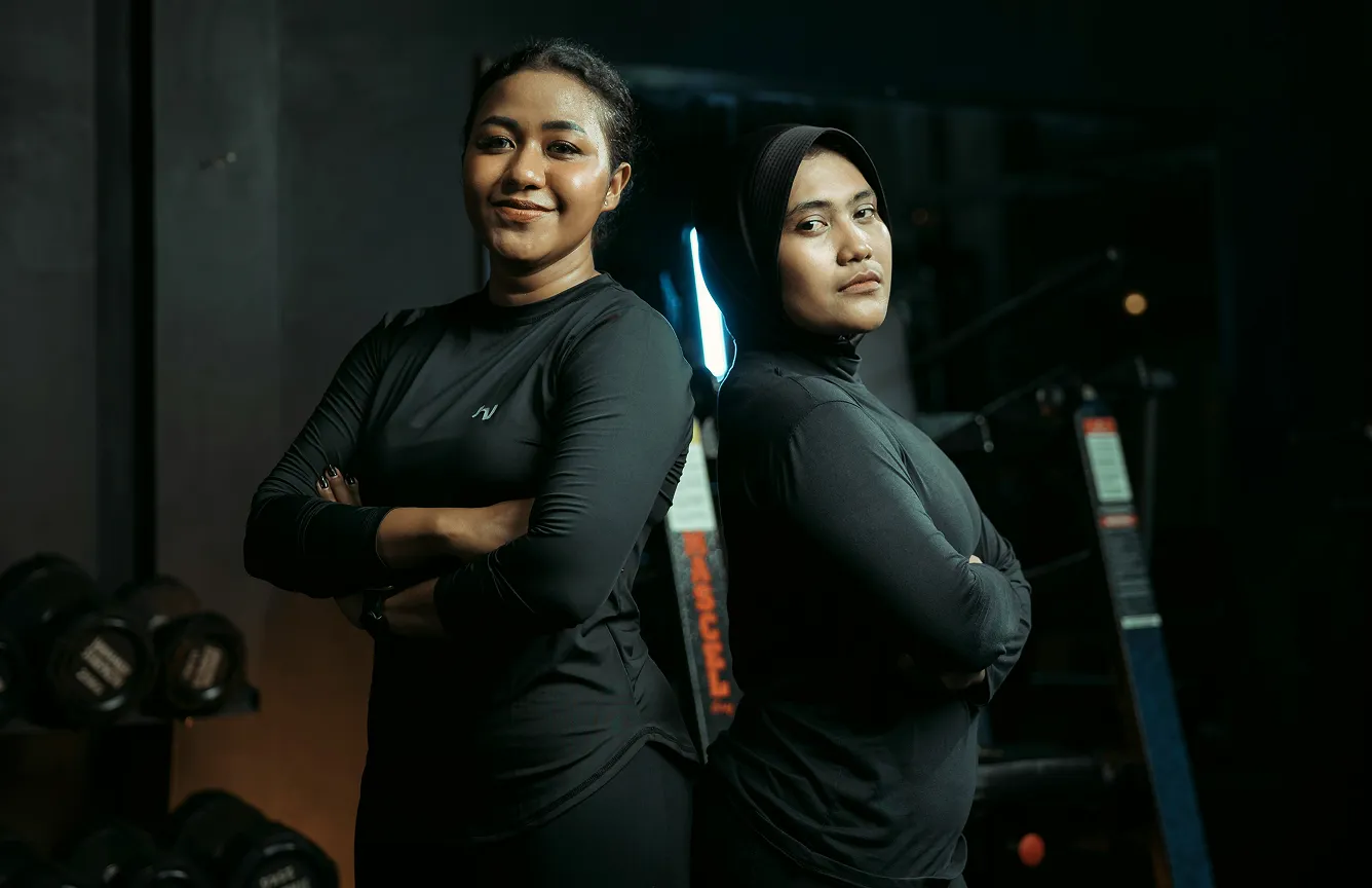 Two women standing back to back in a gym, arms crossed, wearing black workout outfits.
