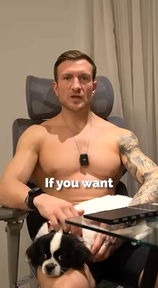 Shirtless man sitting at desk with dog on lap, speaking to camera.