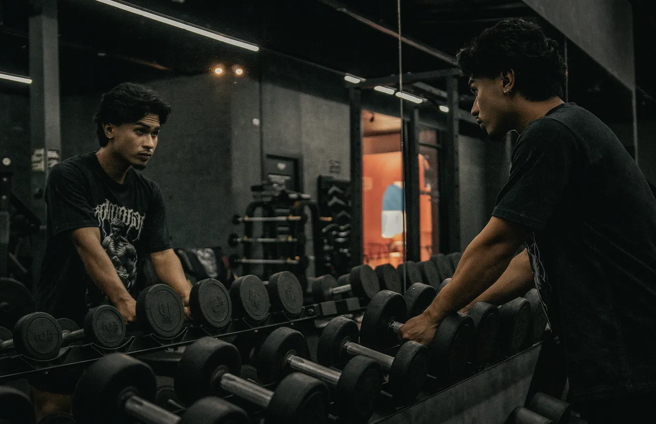 Man lifting dumbbells in a gym while looking at his reflection in a mirror