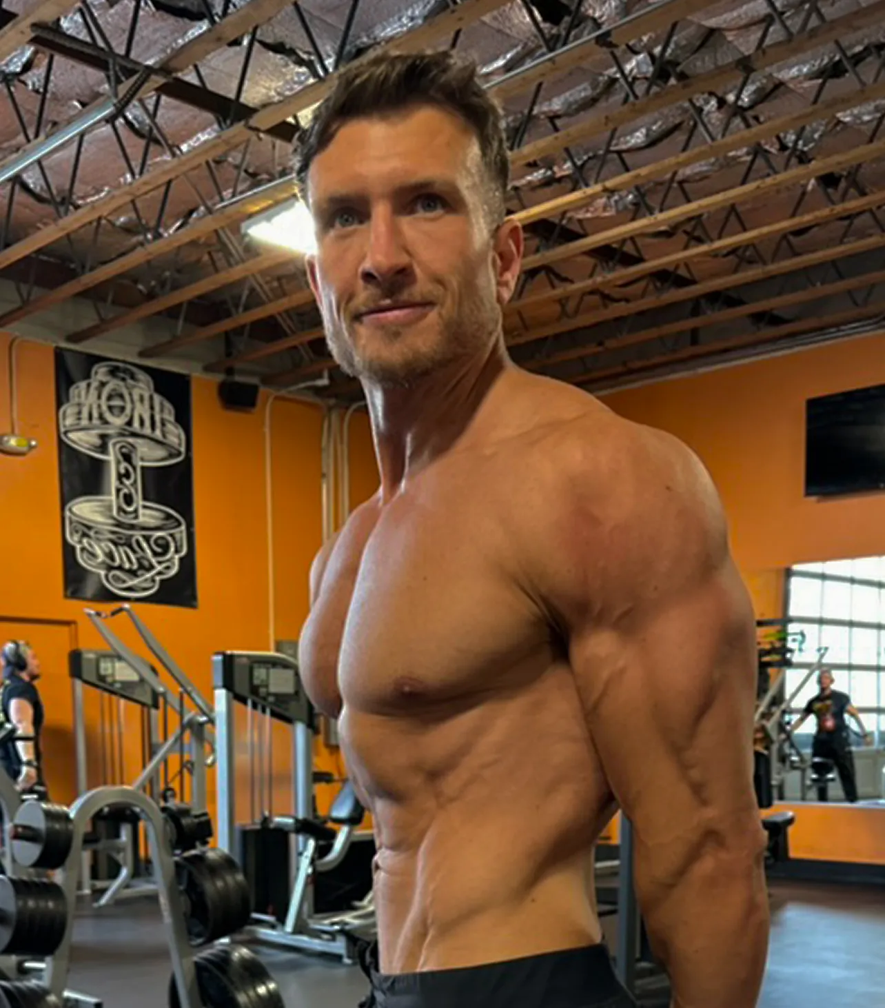 Muscular shirtless man posing in a gym with weightlifting equipment and mirrors in the background.