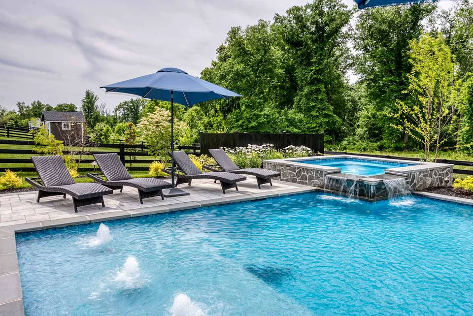 Backyard pool with stone spa, waterfalls, lounge chairs, and blue umbrella in lush garden setting by Deckscapes of Virginia