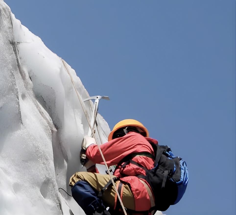 Climber wearing a helmet and backpack ascending an icy mountain face with an ice axe and rope under a clear blue sky.