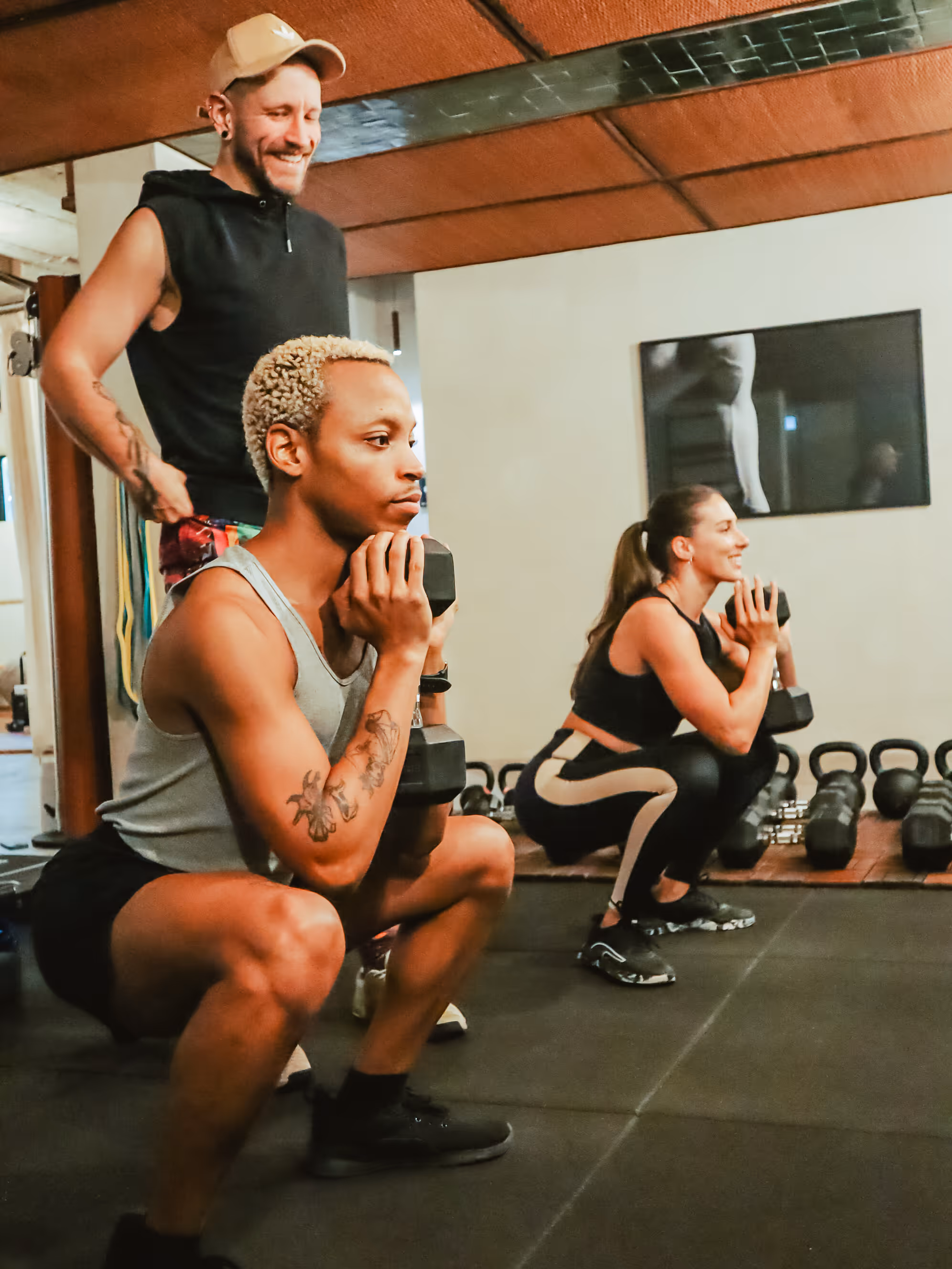Two people squatting with dumbbells in a gym while a trainer watches and smiles.