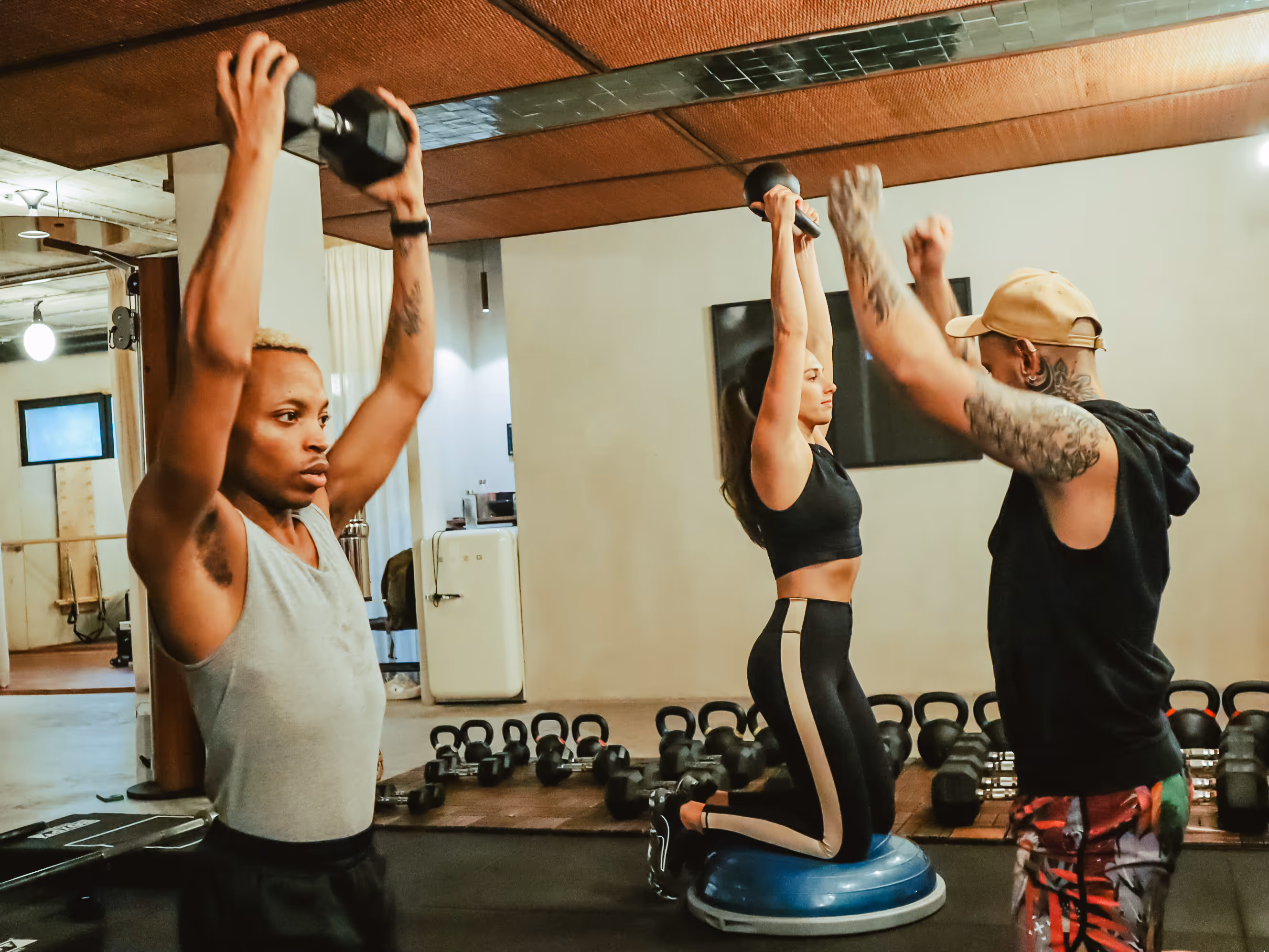 Two people lifting dumbbells overhead while a trainer coaches a woman on a balance trainer.