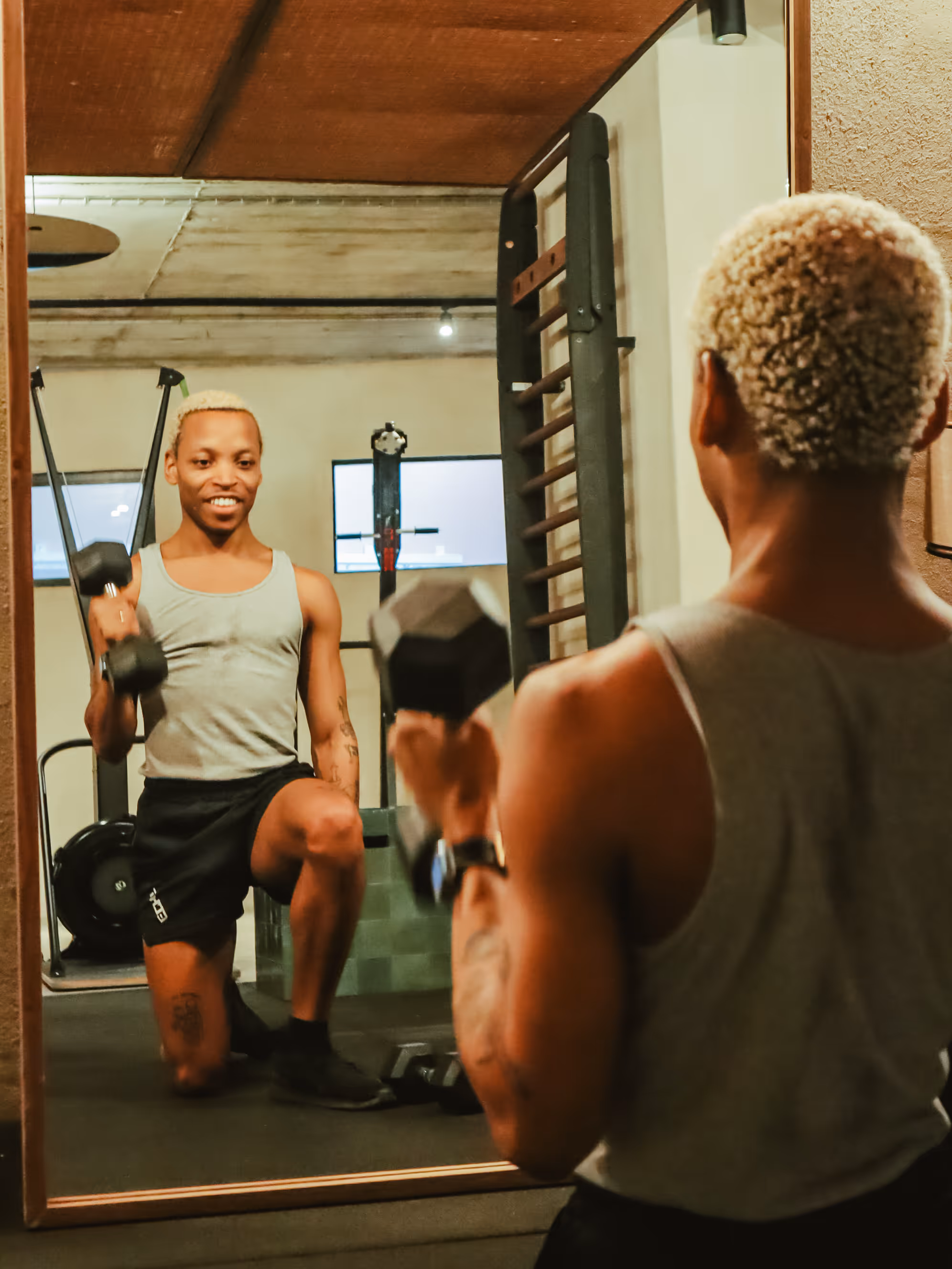 Man with blonde hair kneeling and lifting dumbbells while looking at himself in a gym mirror.