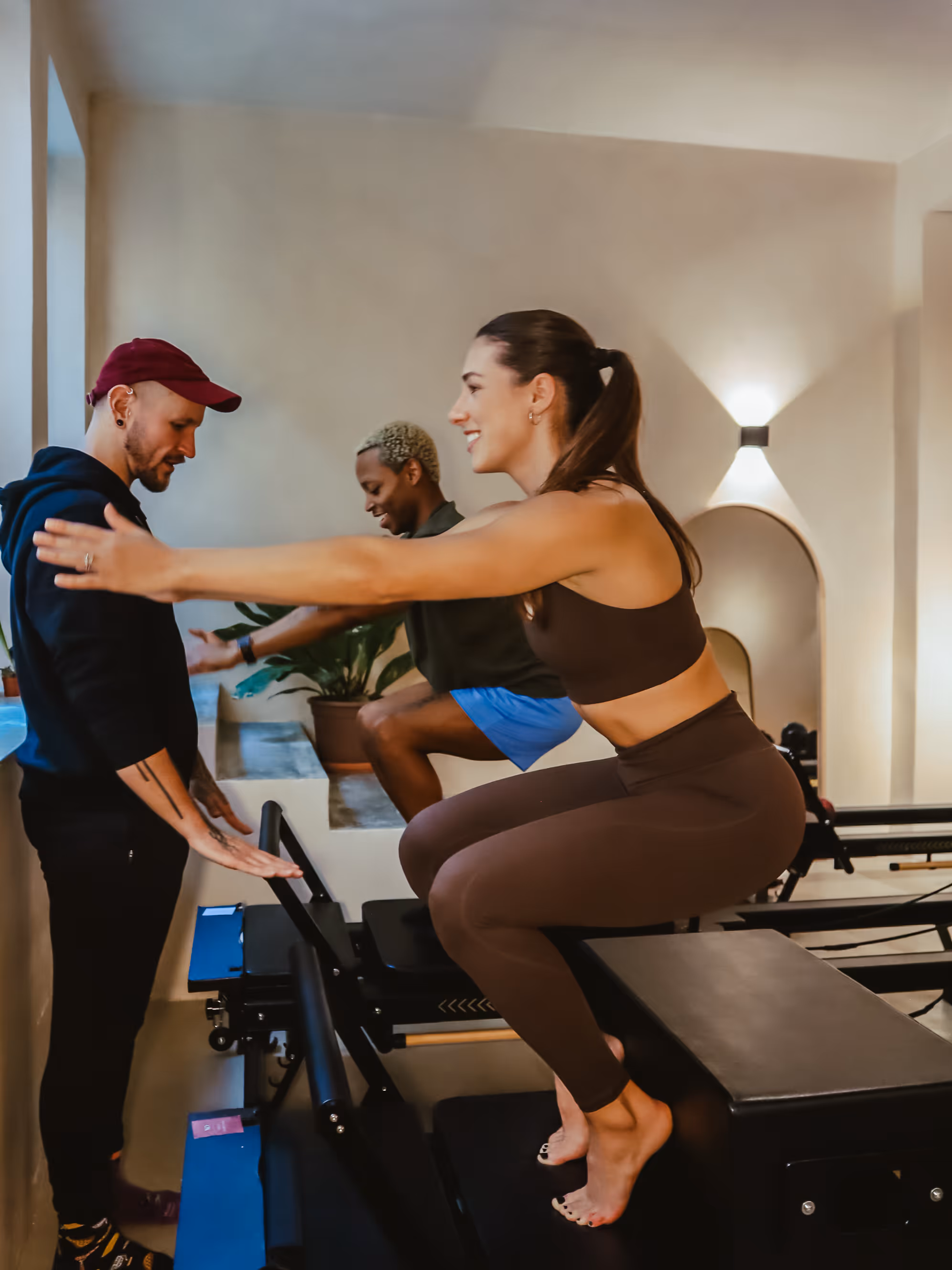 Woman doing squats on Pilates reformer machine while instructor and another person watch and encourage.