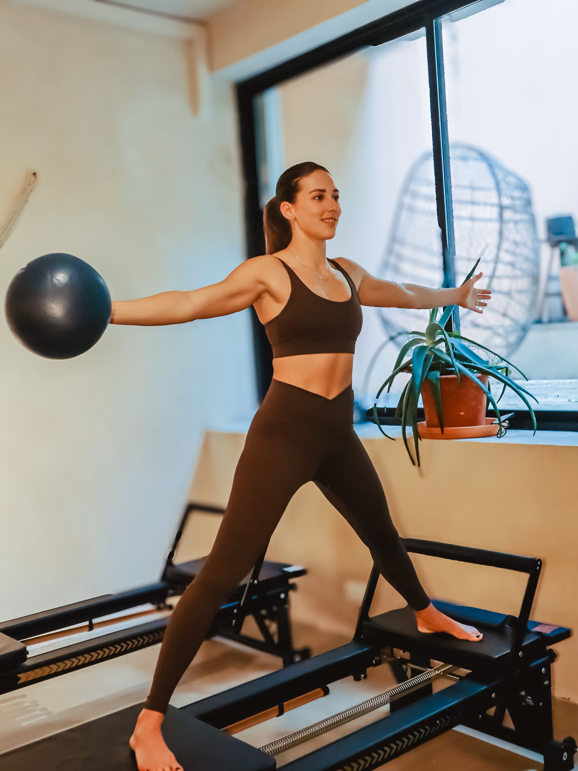Woman in workout clothes holding exercise ball with arms extended, balancing on Pilates reformer machine.