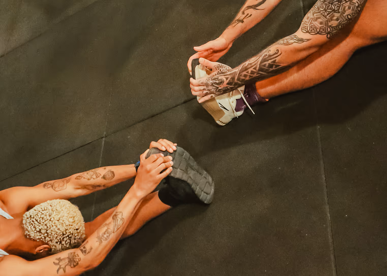 Two tattooed individuals stretching forward to touch their feet on a dark gym floor.