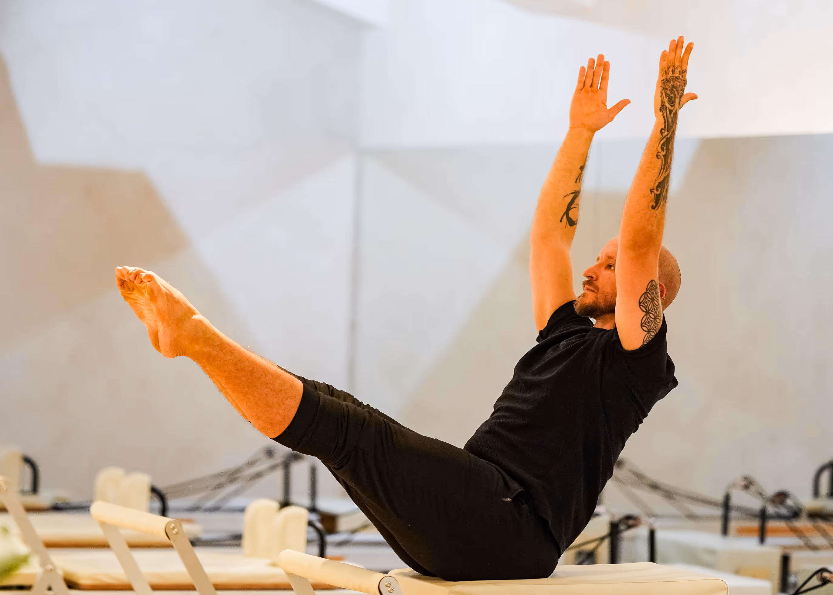 Tattooed man in black workout clothes raising legs and arms while balancing on exercise bench indoors.
