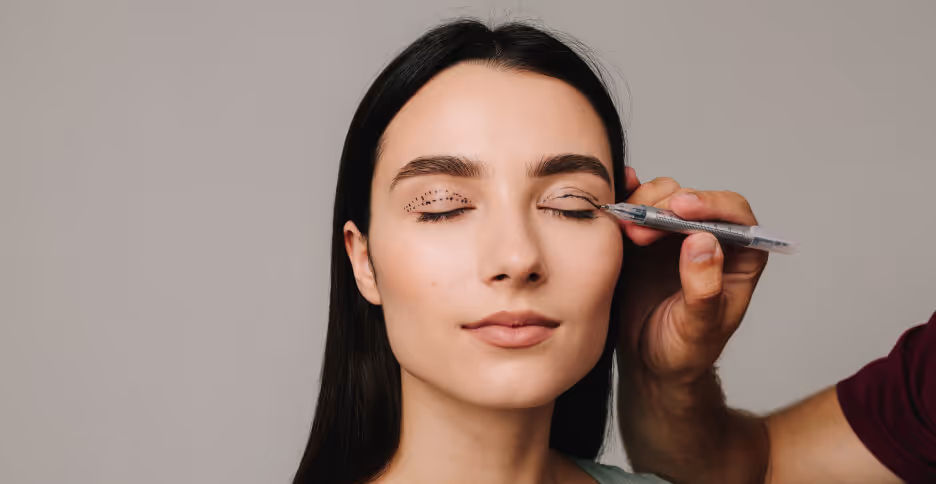 Close-up of a woman with eyes closed having dotted markings drawn on her eyelids by a person holding a marker.