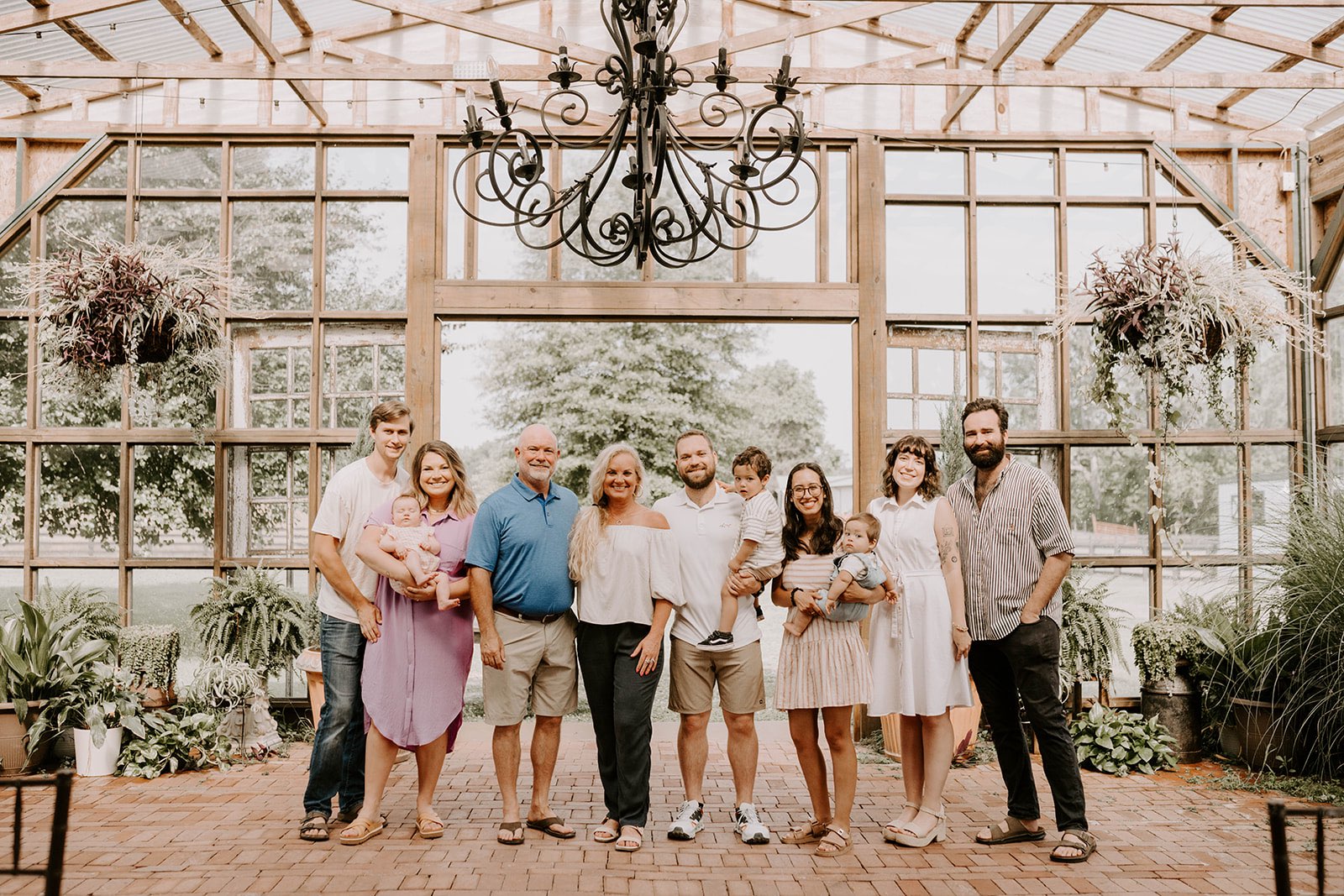 A multi-generational family of nine, including three babies, poses smiling inside a sunlit greenhouse with hanging plants and large windows.