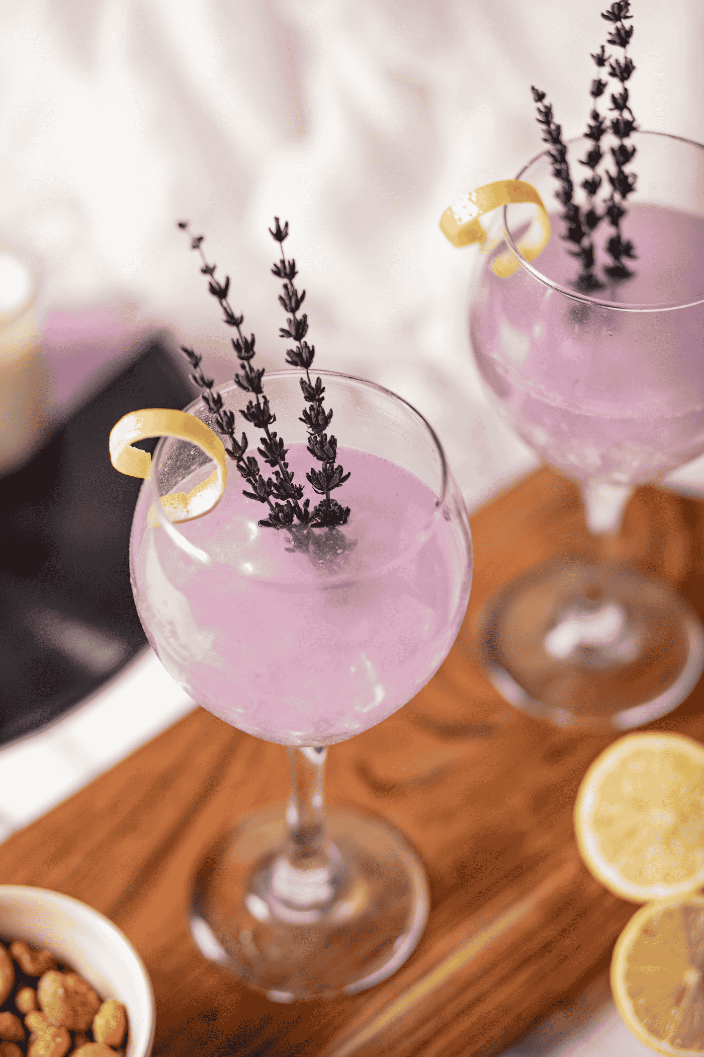 Two glasses of purple cocktail garnished with lemon peel and lavender sprigs on a wooden board with lemon slices and nuts nearby.