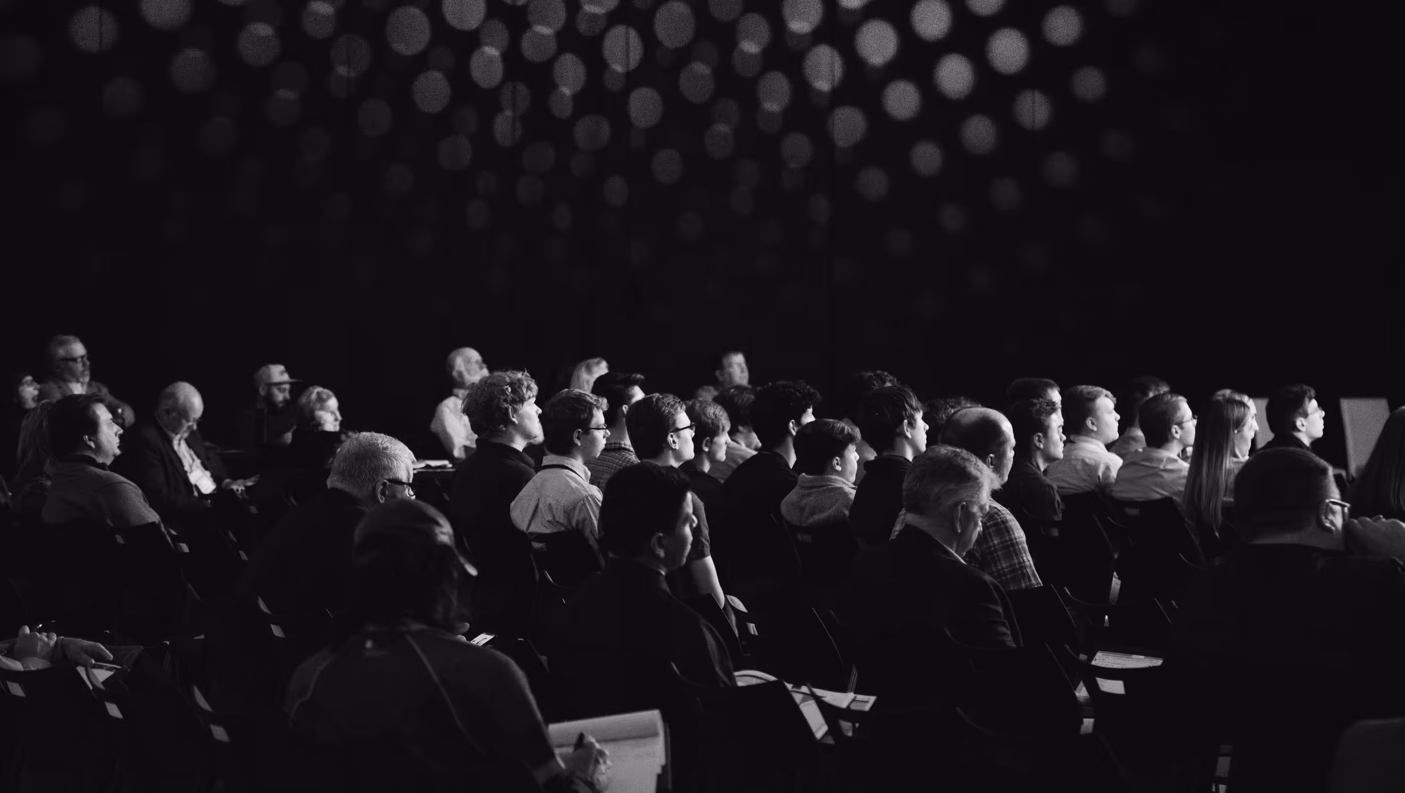 Black and white photo of an audience seated in rows, attentively facing forward, with circular lights visible in the background.