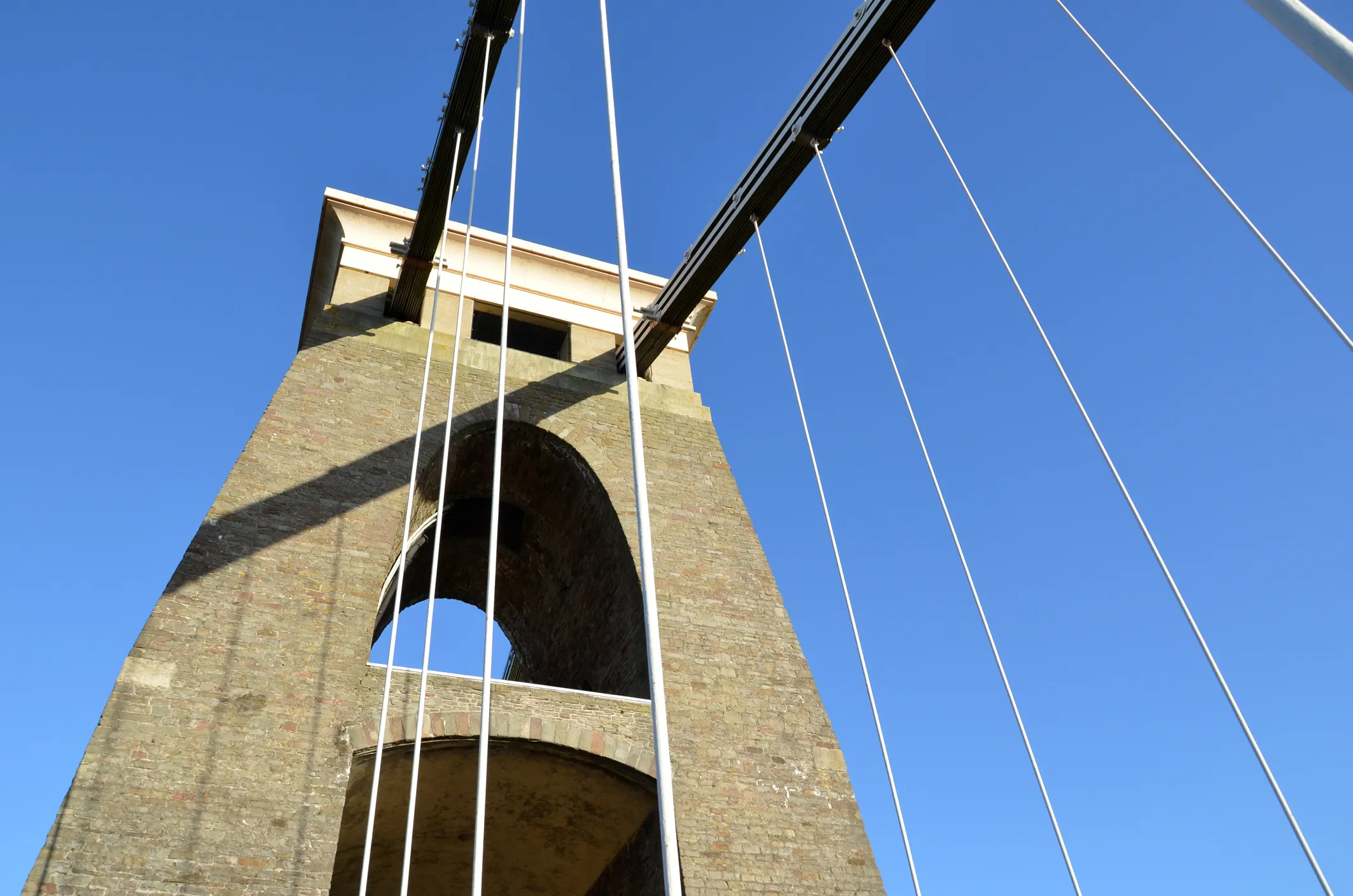 Upward view of a stone suspension bridge tower with white suspension cables against a clear blue sky.