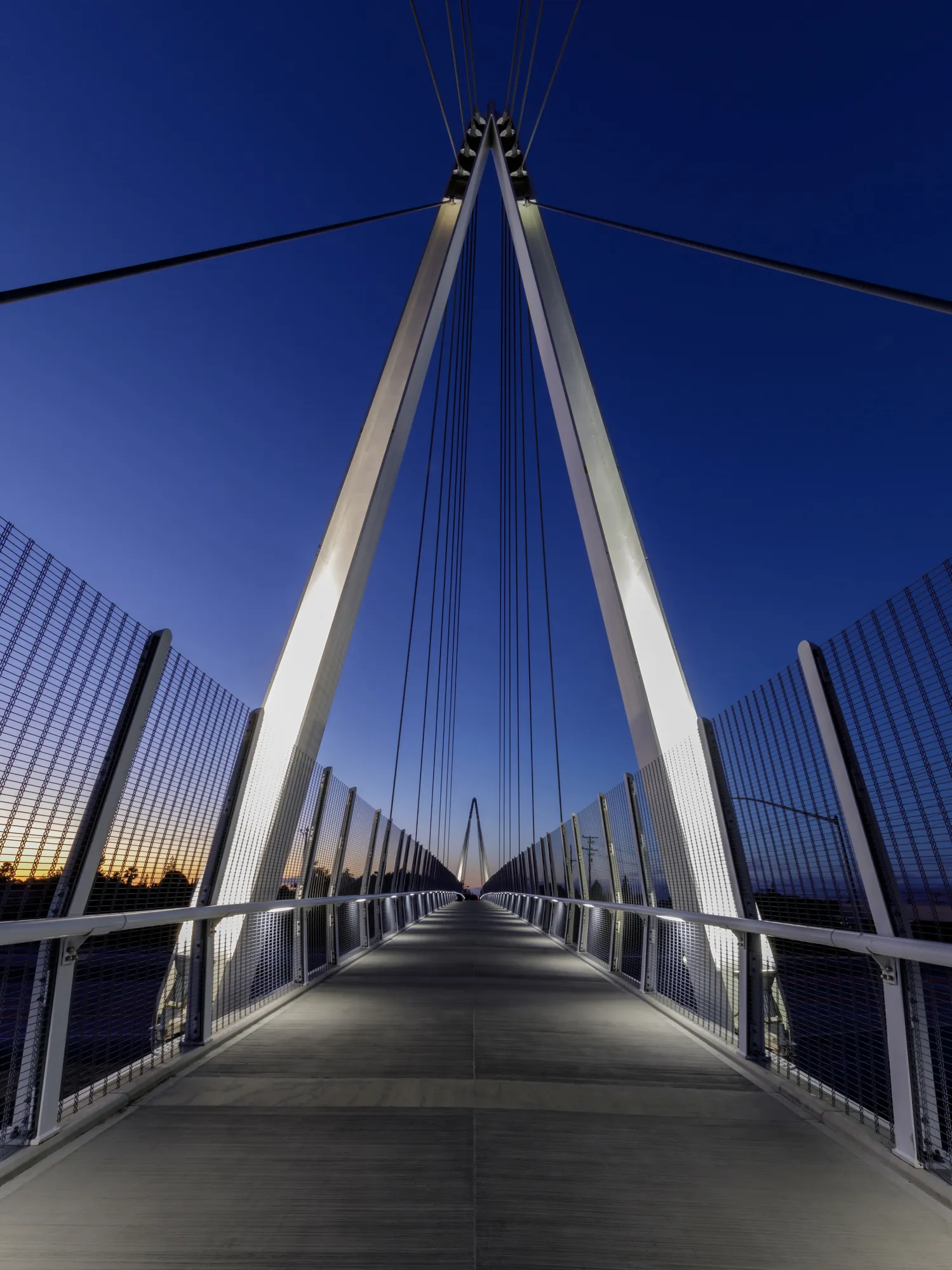 Illuminated modern pedestrian suspension bridge at dusk with clear blue sky.