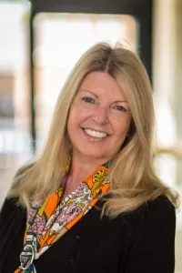 Smiling woman with long blonde hair wearing a black top and colorful scarf, standing indoors in soft natural light.