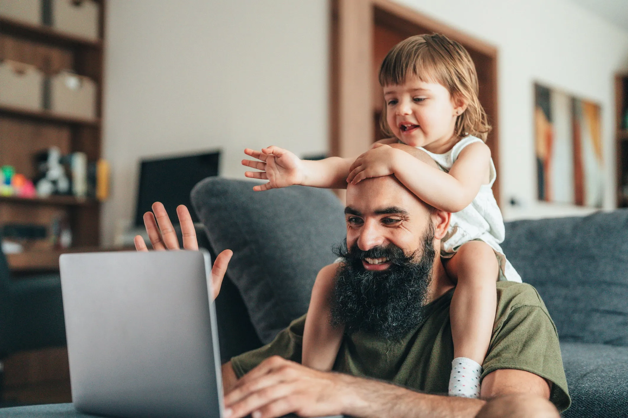 Bearded man smiling and using a laptop with a young child sitting on his shoulders reaching forward.