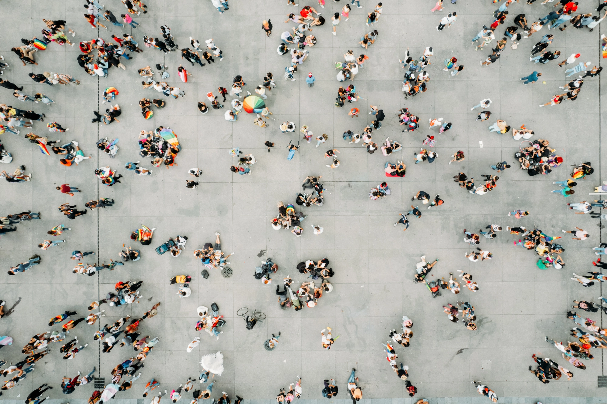 Aerial view of a large crowd of people gathered on a gray concrete surface, many wearing colorful clothing and holding rainbow flags and umbrellas.