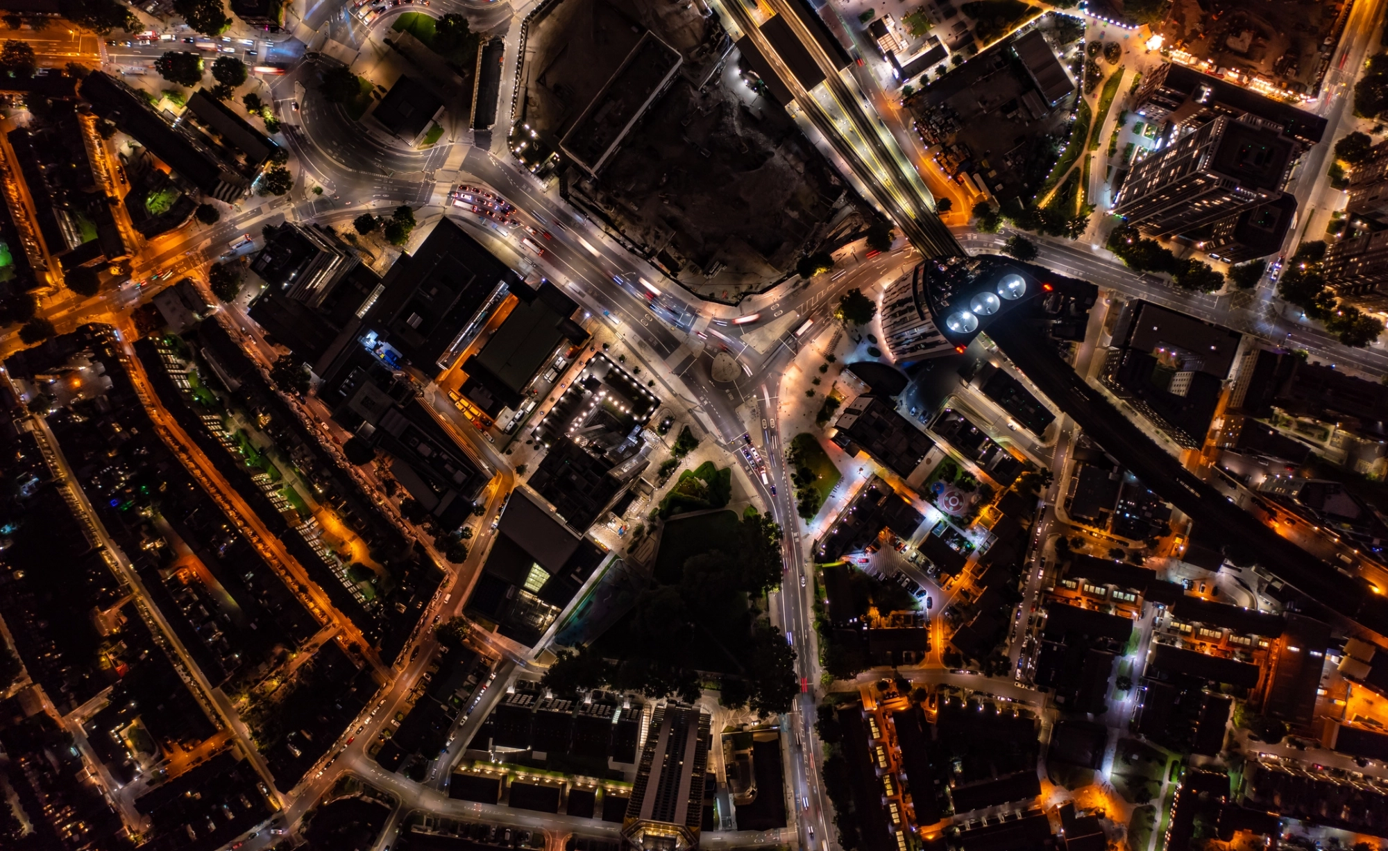 Aerial night view of a city intersection with illuminated streets, buildings, and moving vehicles.
