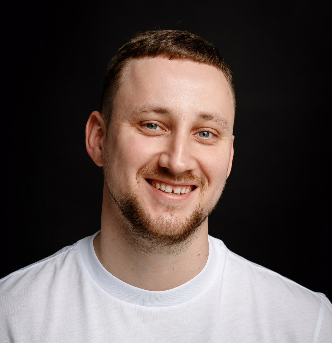 Smiling man with short brown hair and beard wearing a white t-shirt against a black background.
