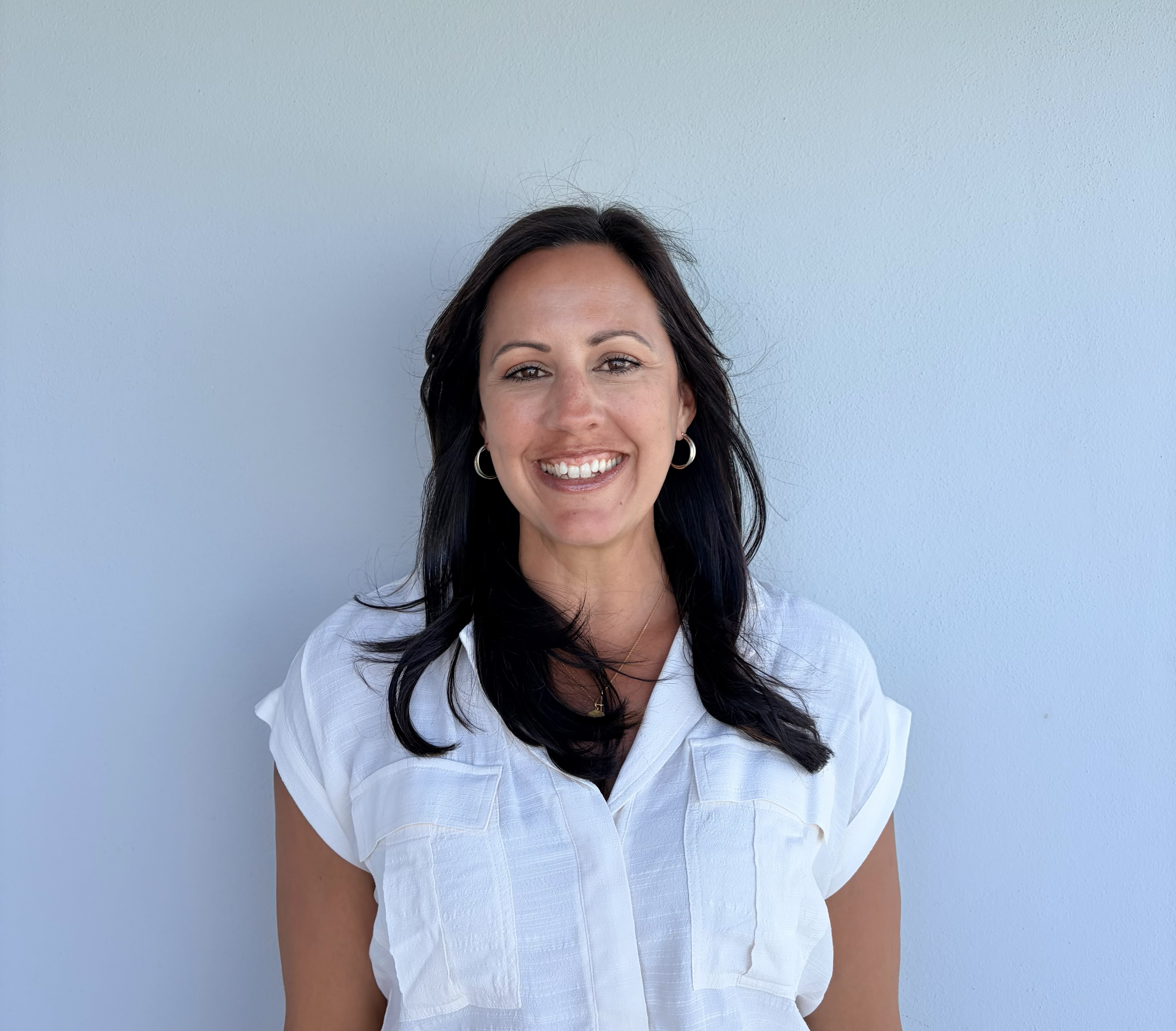 Smiling woman with long dark hair wearing a white shirt standing in front of a pale blue wall.