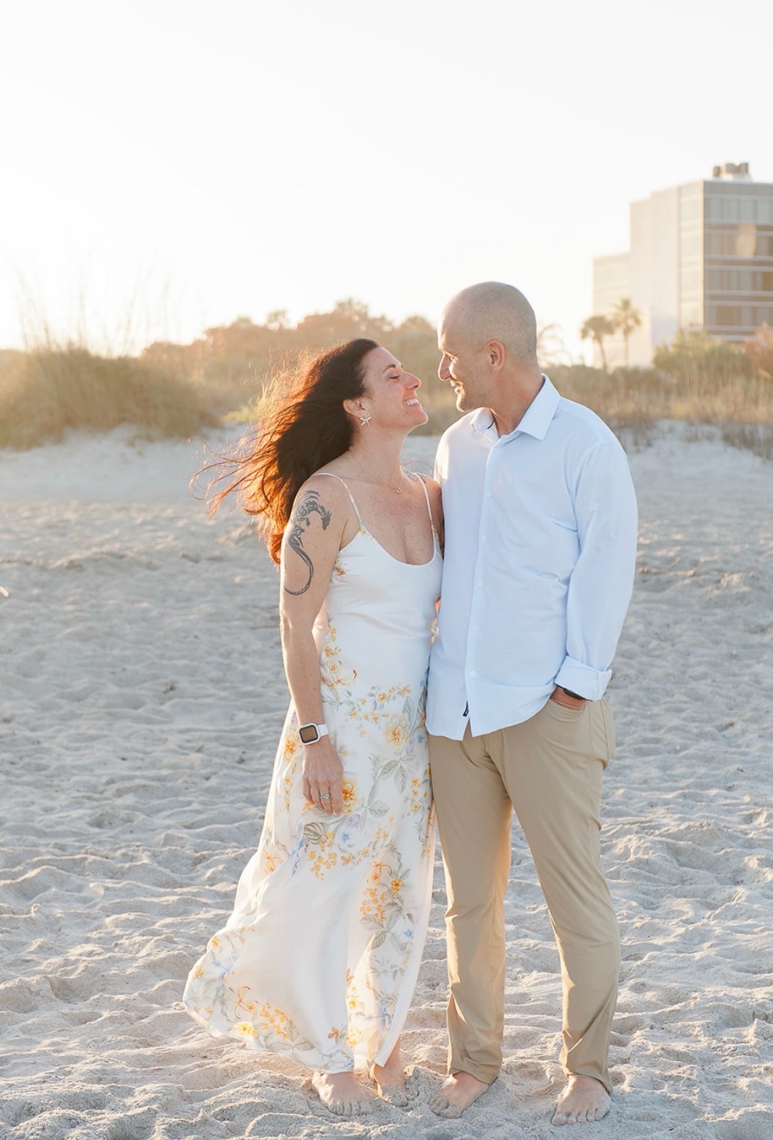 Couple smiling at each other barefoot on a sandy beach at sunset with grassy dunes behind.