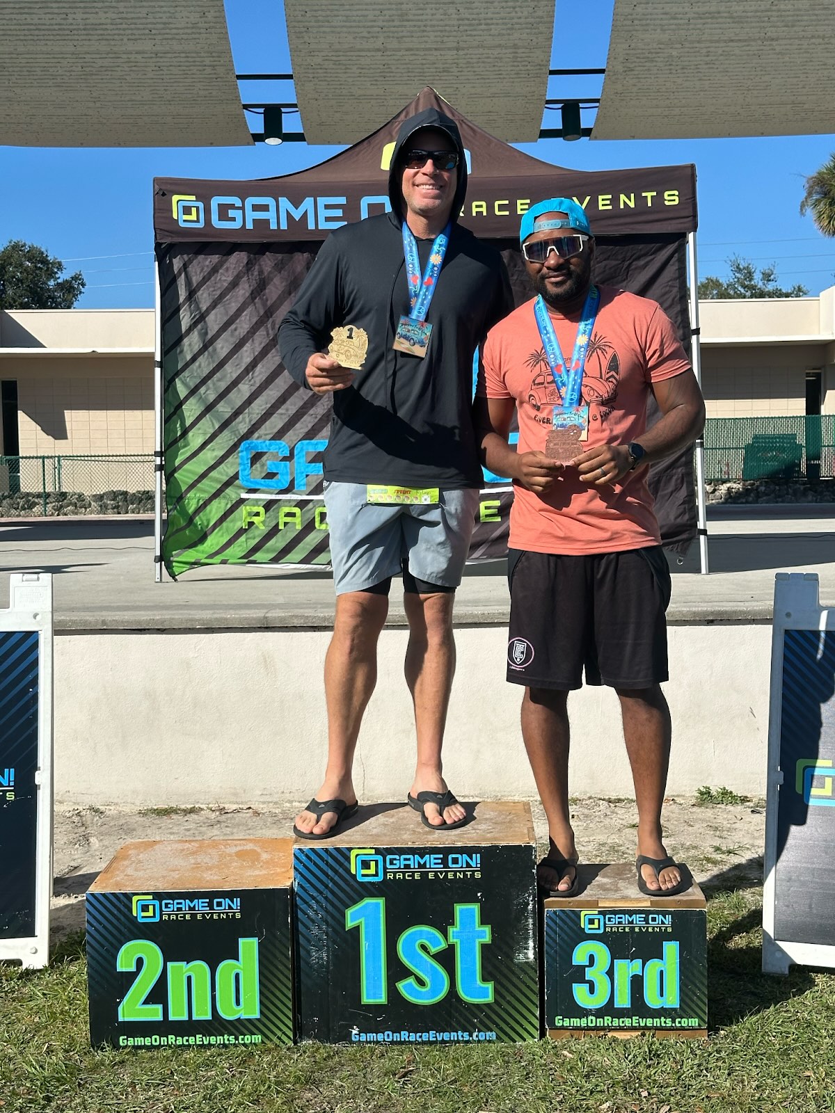 Two men on race award podium holding medals in front of a Game On Race Events banner outdoors.