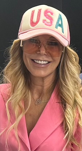Woman smiling wearing USA cap, pink blazer, sunglasses, and a necklace with Bonita pendant.