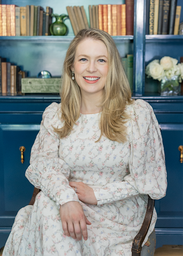 Smiling woman with long blonde hair wearing a white floral dress, seated in front of blue shelves with books and flowers.