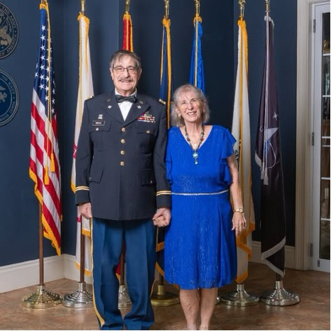 Man in military uniform holding hands with woman in blue dress standing in front of flags.