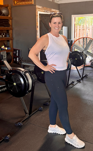 Smiling woman in white tank top and leggings stands beside rowing machines in a gym.