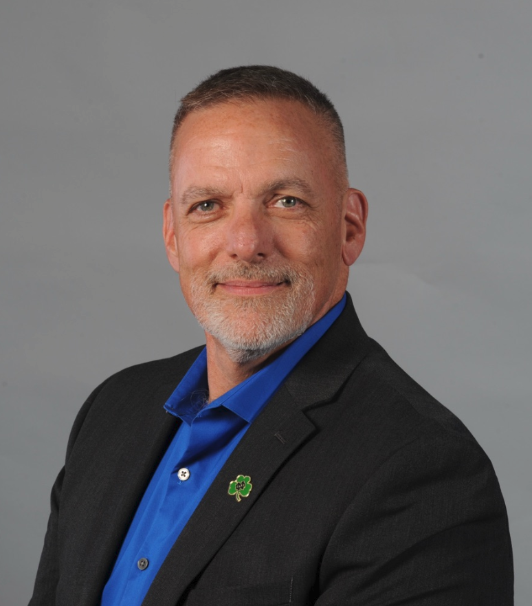 Smiling man with short gray hair and beard wearing a black blazer and blue shirt with a green clover pin.