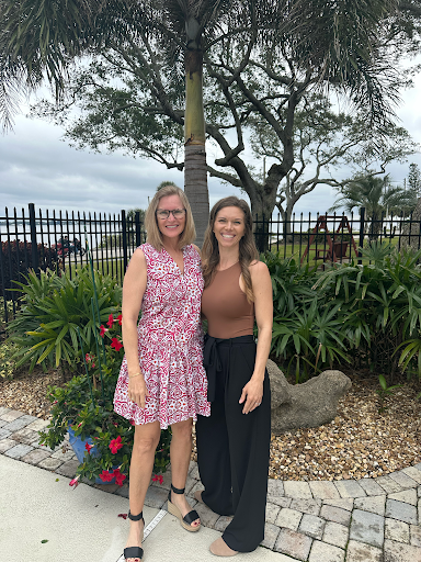 Two smiling women standing near plants and a palm tree, with a black fence and cloudy sky behind them.