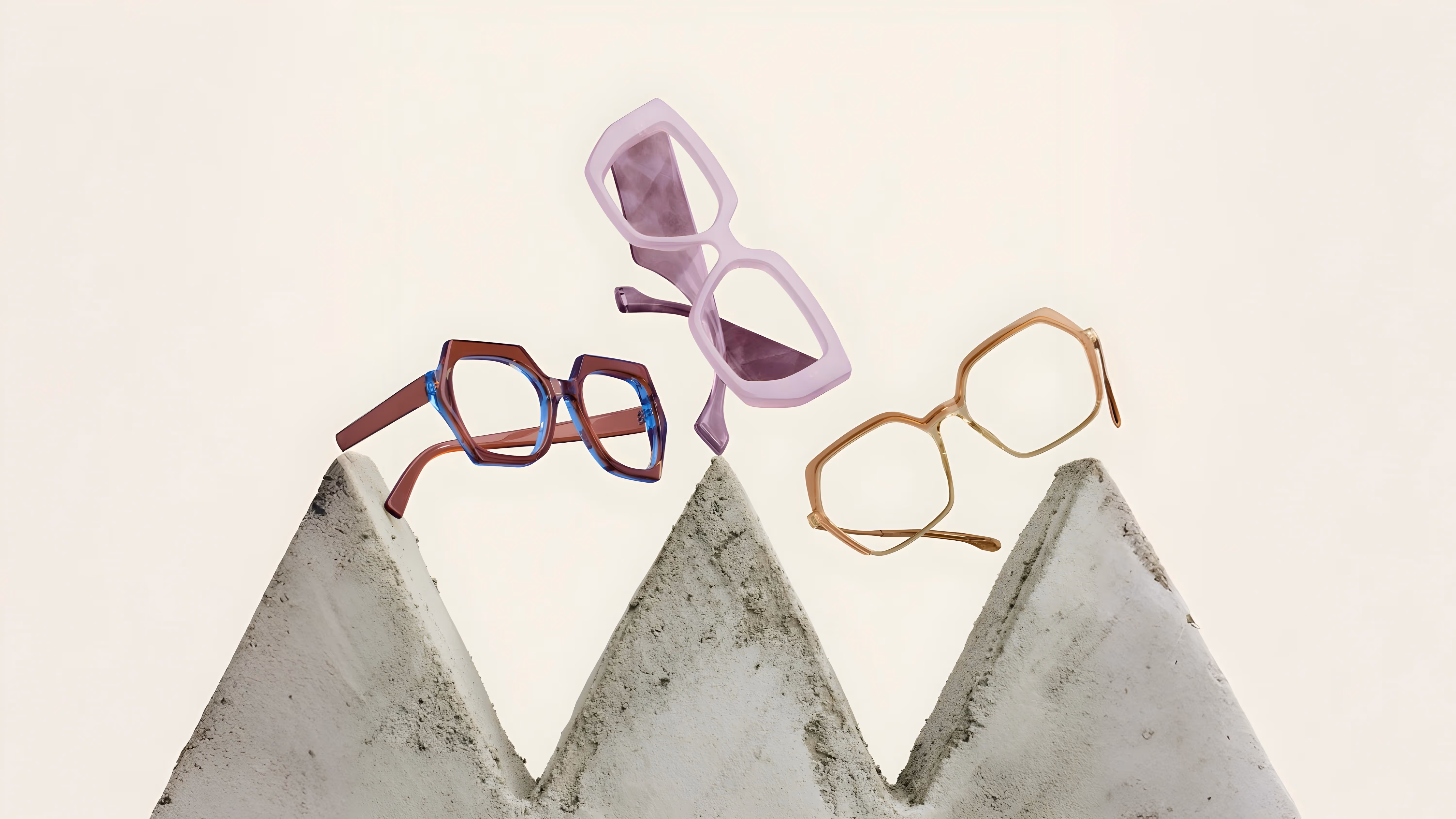 Three pairs of modern eyeglasses in translucent blue, purple, and beige balanced on pointed concrete blocks against a plain background.