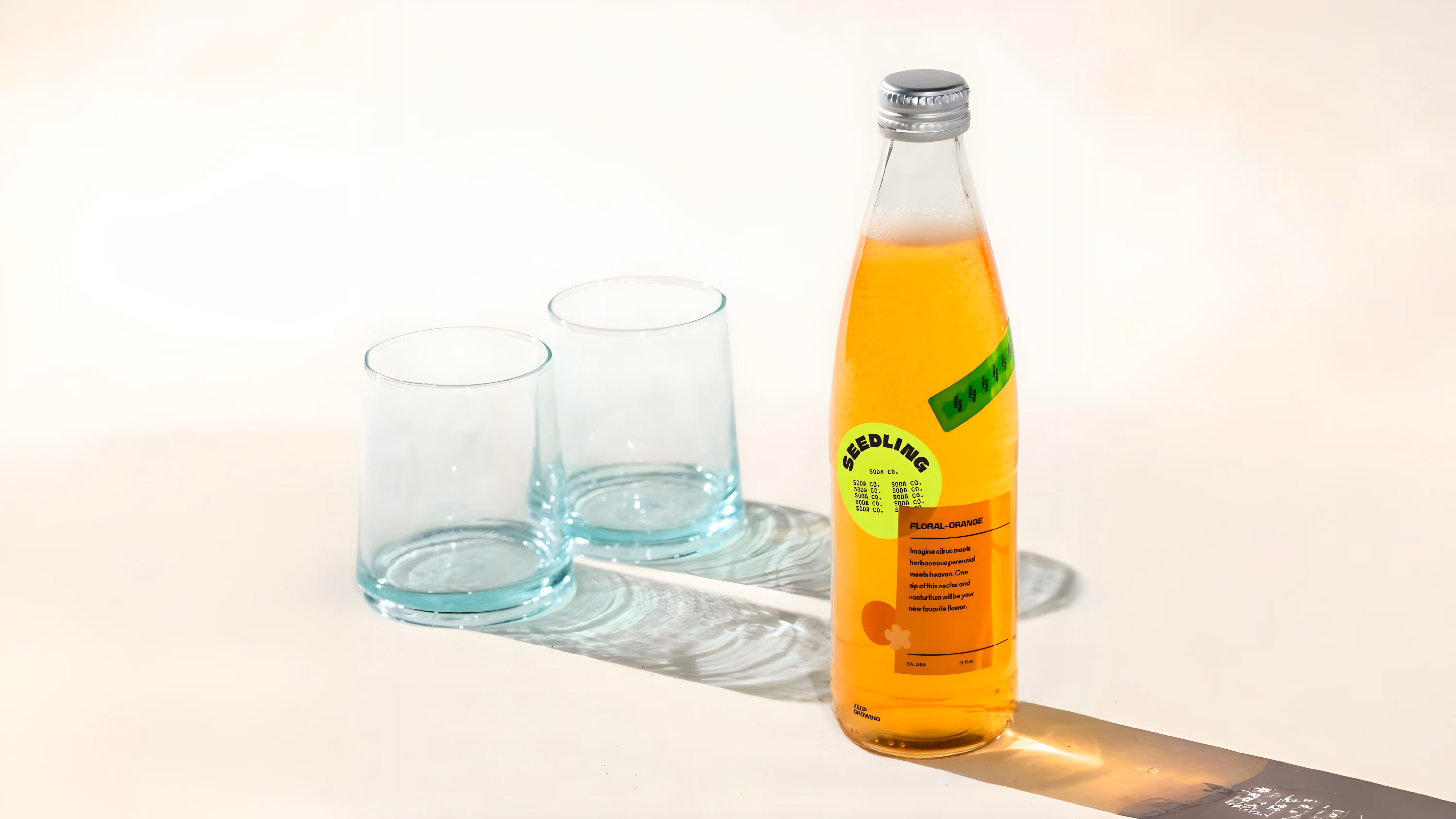 Glass bottle of orange floral soda labeled 'Seedling' next to two empty clear glasses on a white surface.