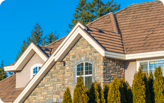 Close-up view of a house roof with brown shingles, stone and stucco exterior walls, and tall green shrubs in front under a clear blue sky.