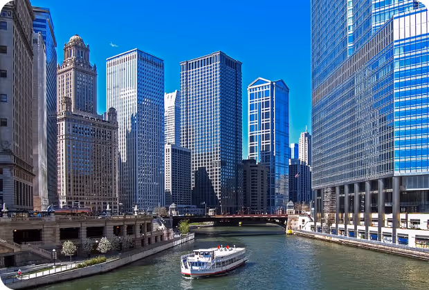 Boat cruising on a river flanked by tall skyscrapers under a clear blue sky in a city.