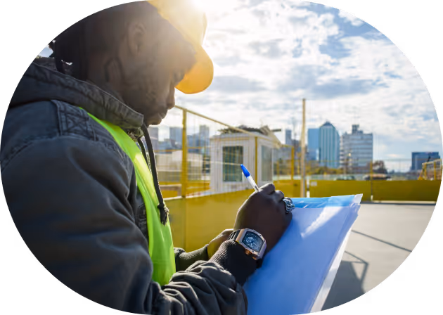 Construction worker wearing a yellow hard hat and reflective vest writing on a clipboard with city buildings in the background.