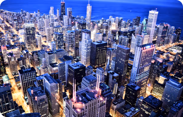 Aerial nighttime view of a cityscape with illuminated skyscrapers and streets leading to a large body of water.
