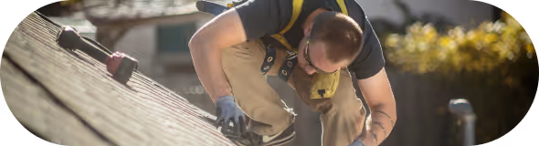 Roofer wearing gloves and safety harness working on a sloped roof under sunlight.