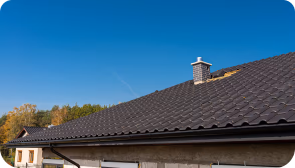 Roof of a house with dark brown tiles and a small chimney under a clear blue sky.