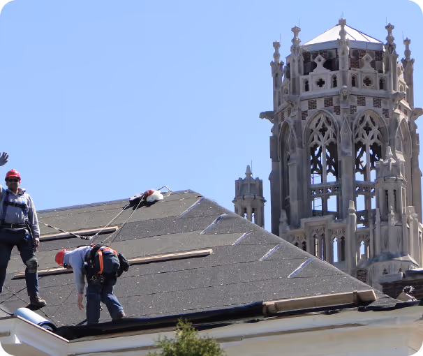 Two construction workers wearing red helmets working on a sloped roof with a large ornate stone tower in the background under a clear blue sky.