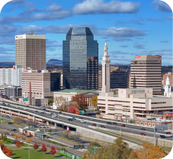Cityscape with mid-rise office buildings, a highway with cars, and colorful autumn trees under a partly cloudy sky.